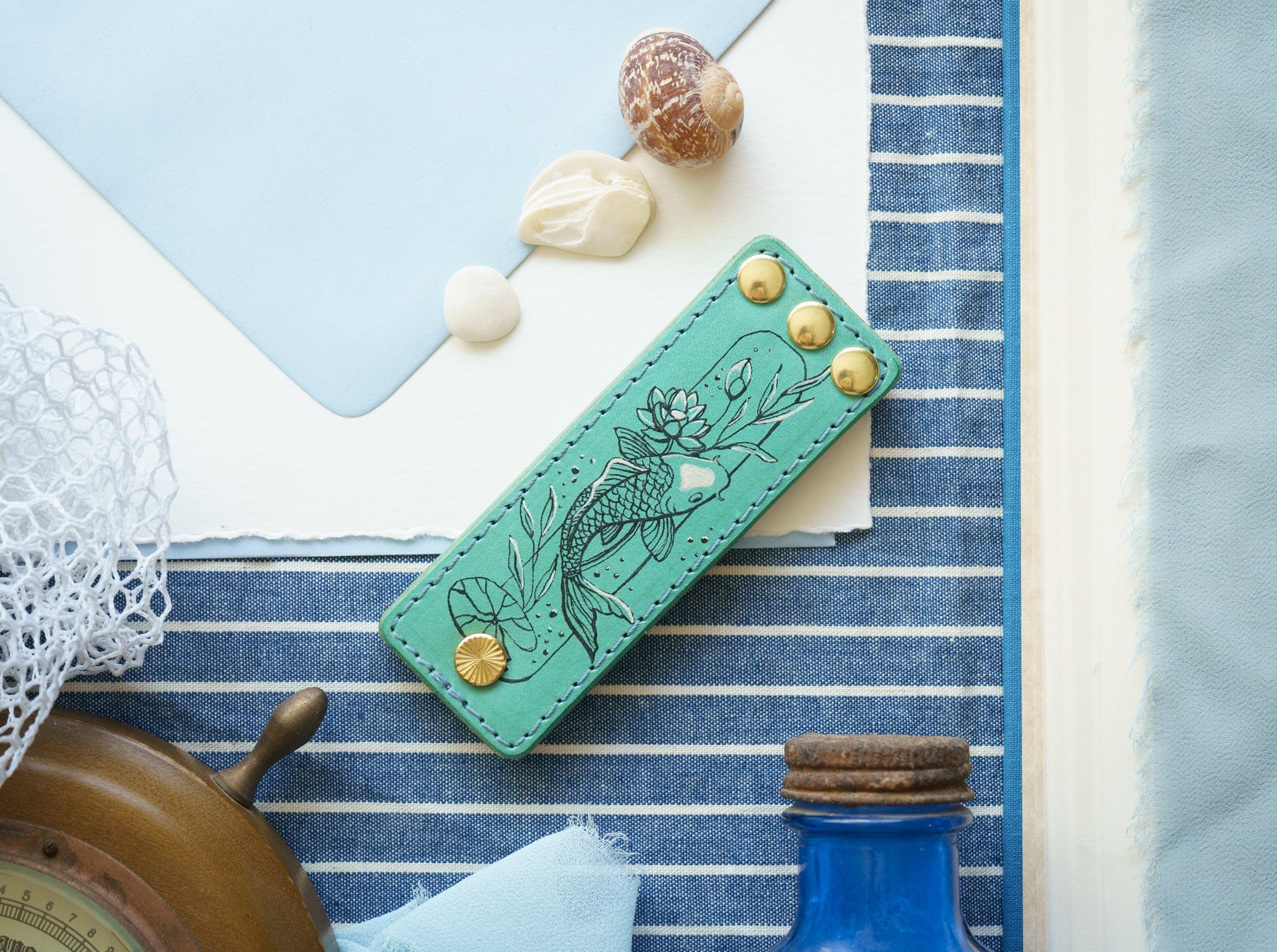 Tattooed blue leather pocket paddle on a flat lay of blue linen and various ocean items.