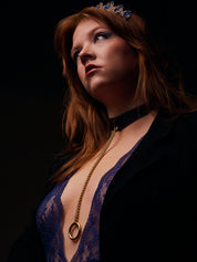 Woman wearing a blue lace top and black blazer with a black leather necklace with gold chain against a dark background
