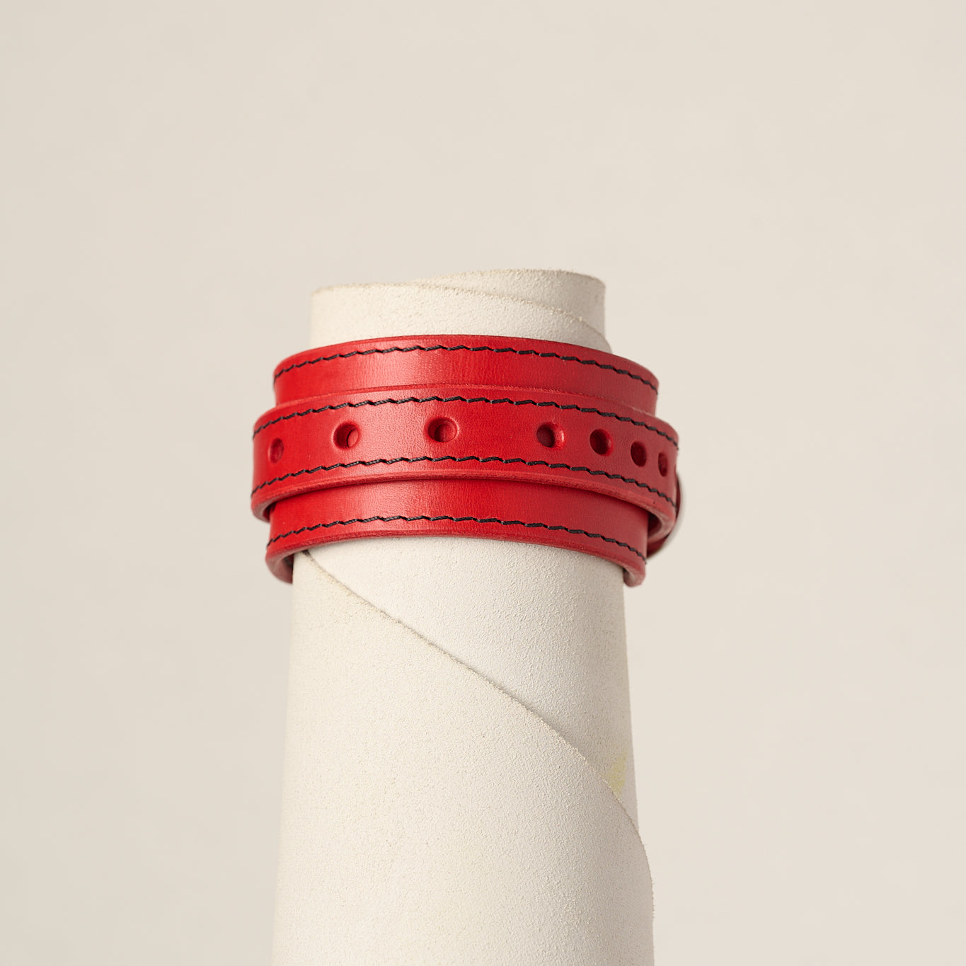 A red leather wrap bracelet displayed on a white cylindrical mannequin stand against a light beige background.