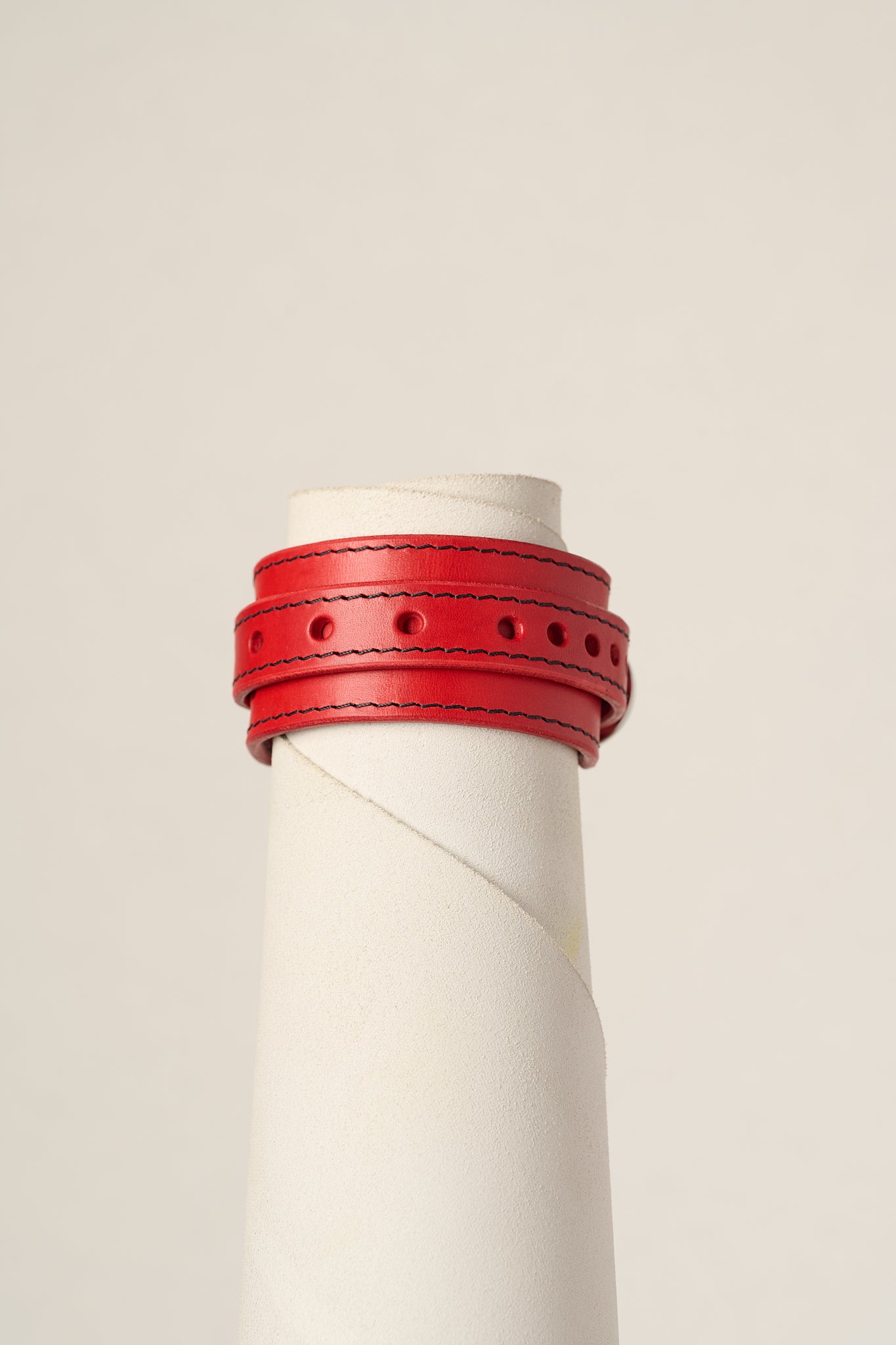A red leather wrap bracelet displayed on a white cylindrical mannequin stand against a light beige background.