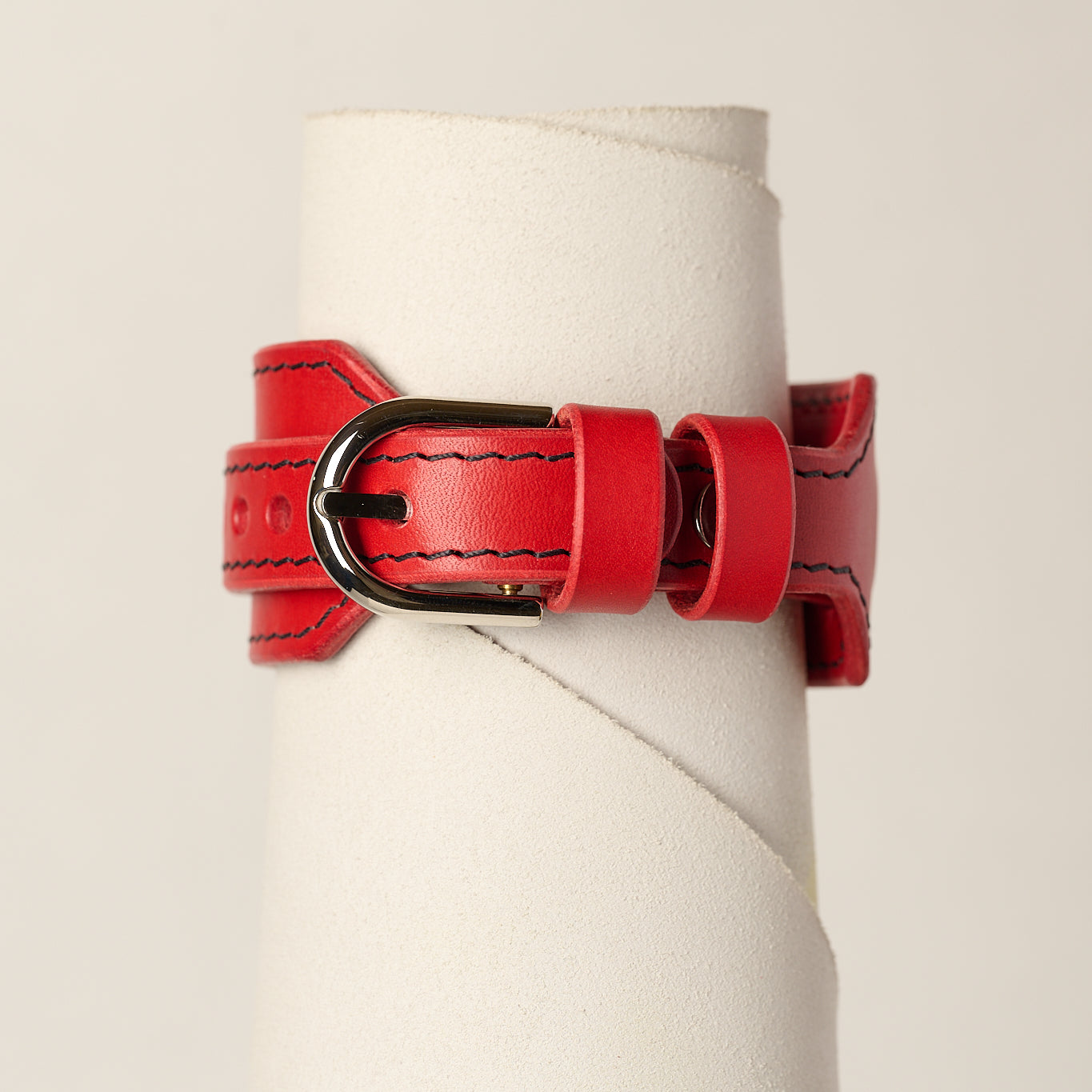 A red leather belt bracelet with black contrast stitching and a black metal buckle, displayed on a white cylindrical stand against a light beige background.