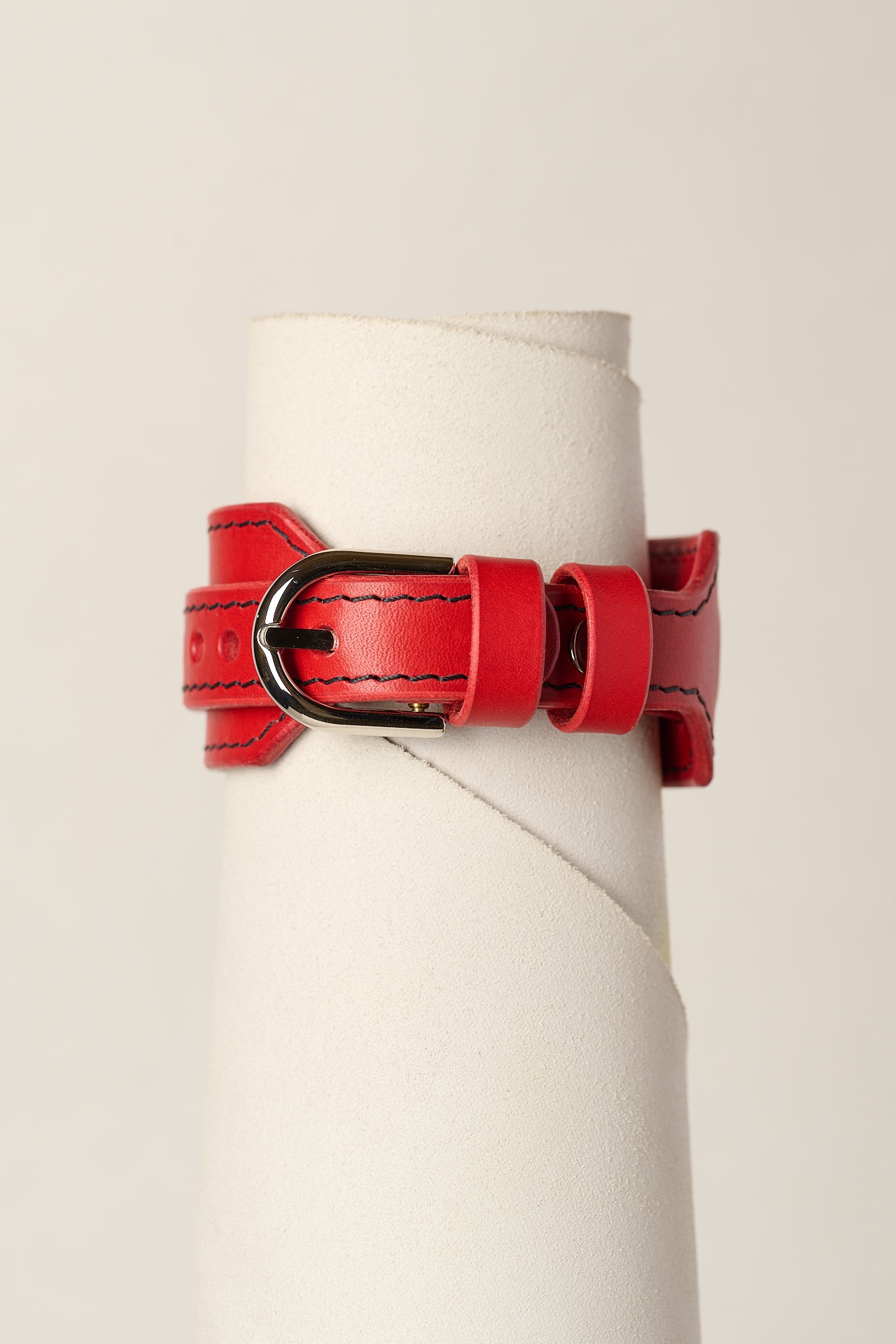 A red leather belt bracelet with black contrast stitching and a black metal buckle, displayed on a white cylindrical stand against a light beige background.