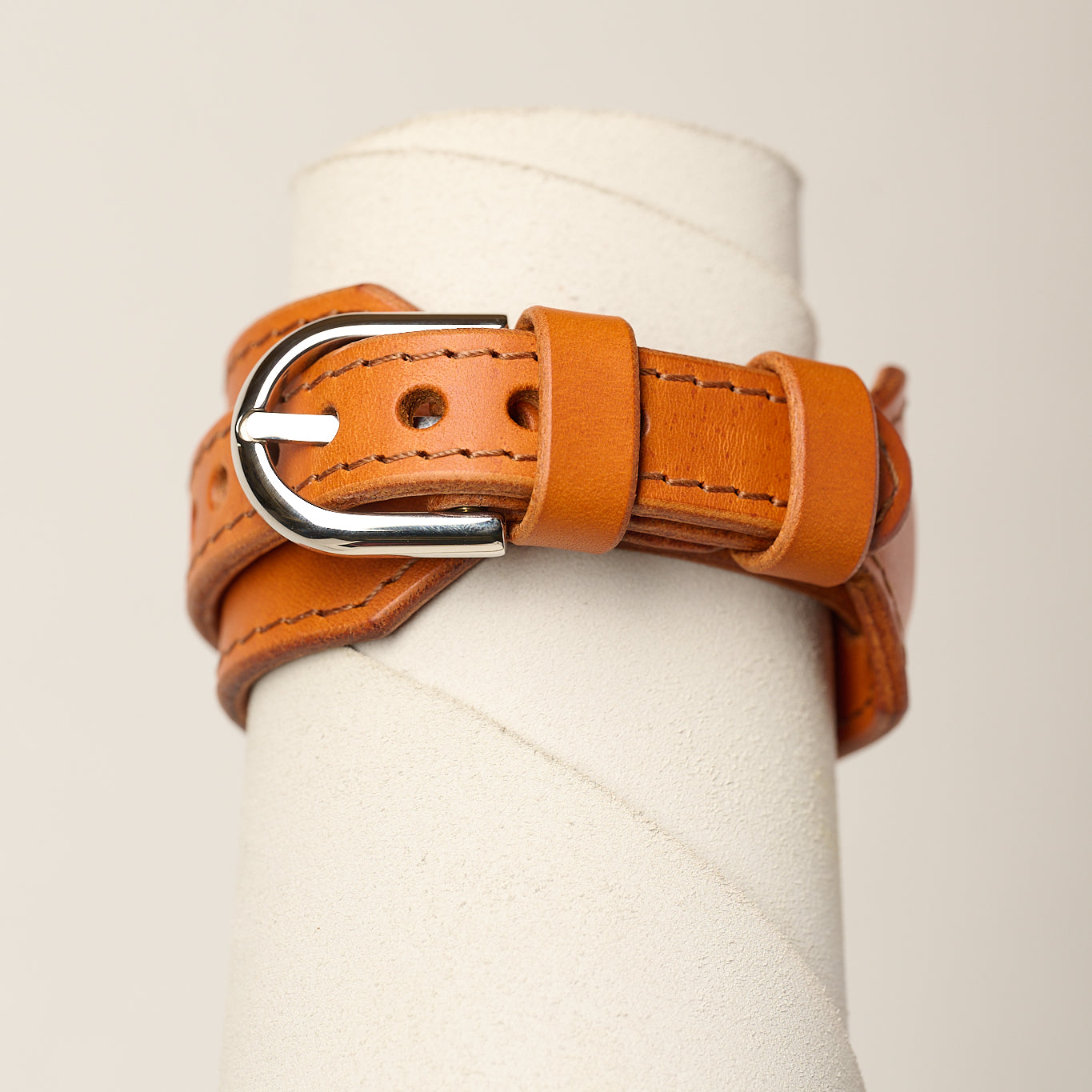 A orange leather belt with a silver metal buckle, wrapped around a white cylindrical display stand against a light beige background.
