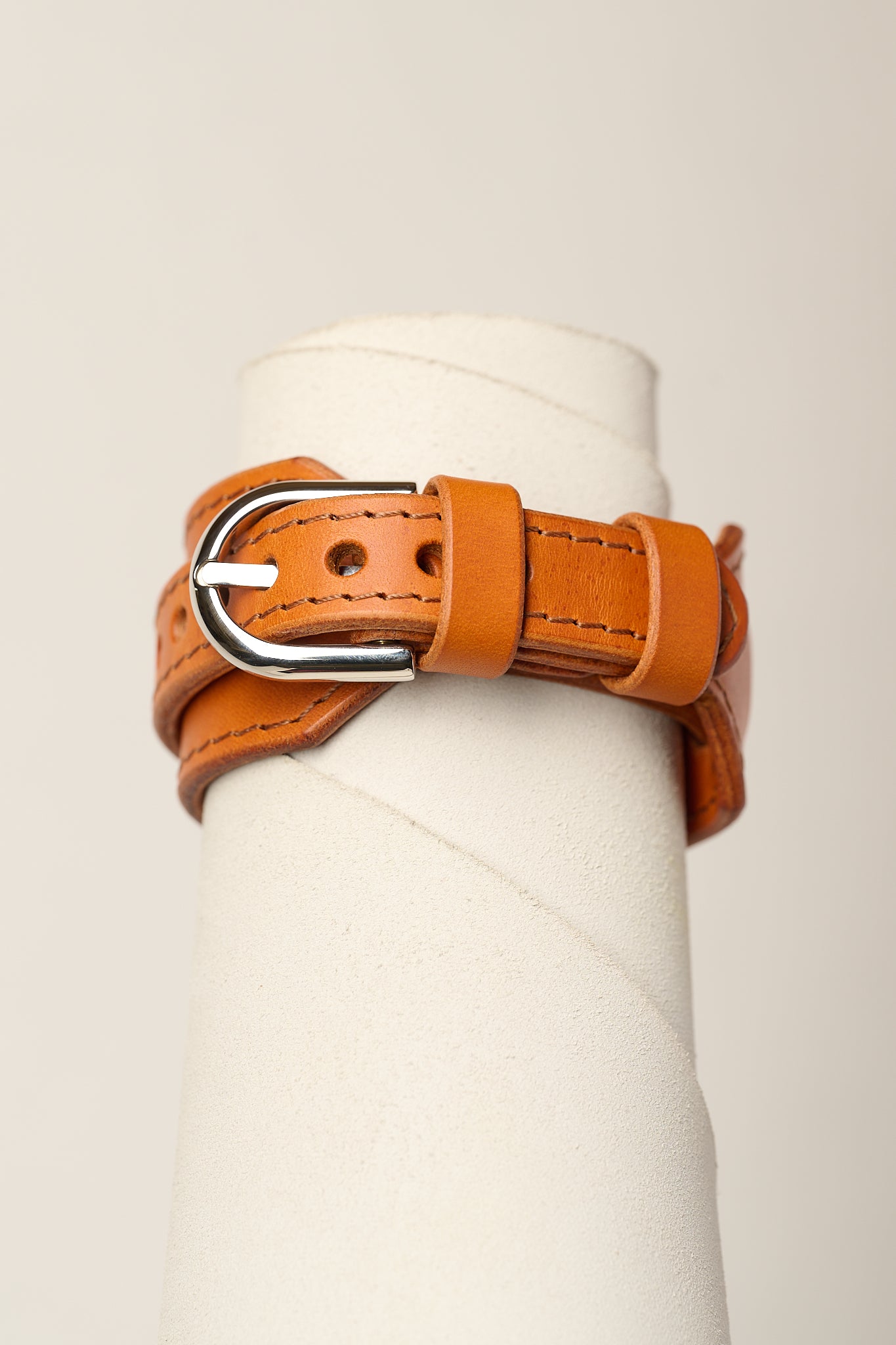 A orange leather belt with a silver metal buckle, wrapped around a white cylindrical display stand against a light beige background.