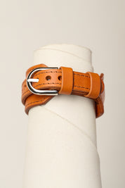 A orange leather belt with a silver metal buckle, wrapped around a white cylindrical display stand against a light beige background.