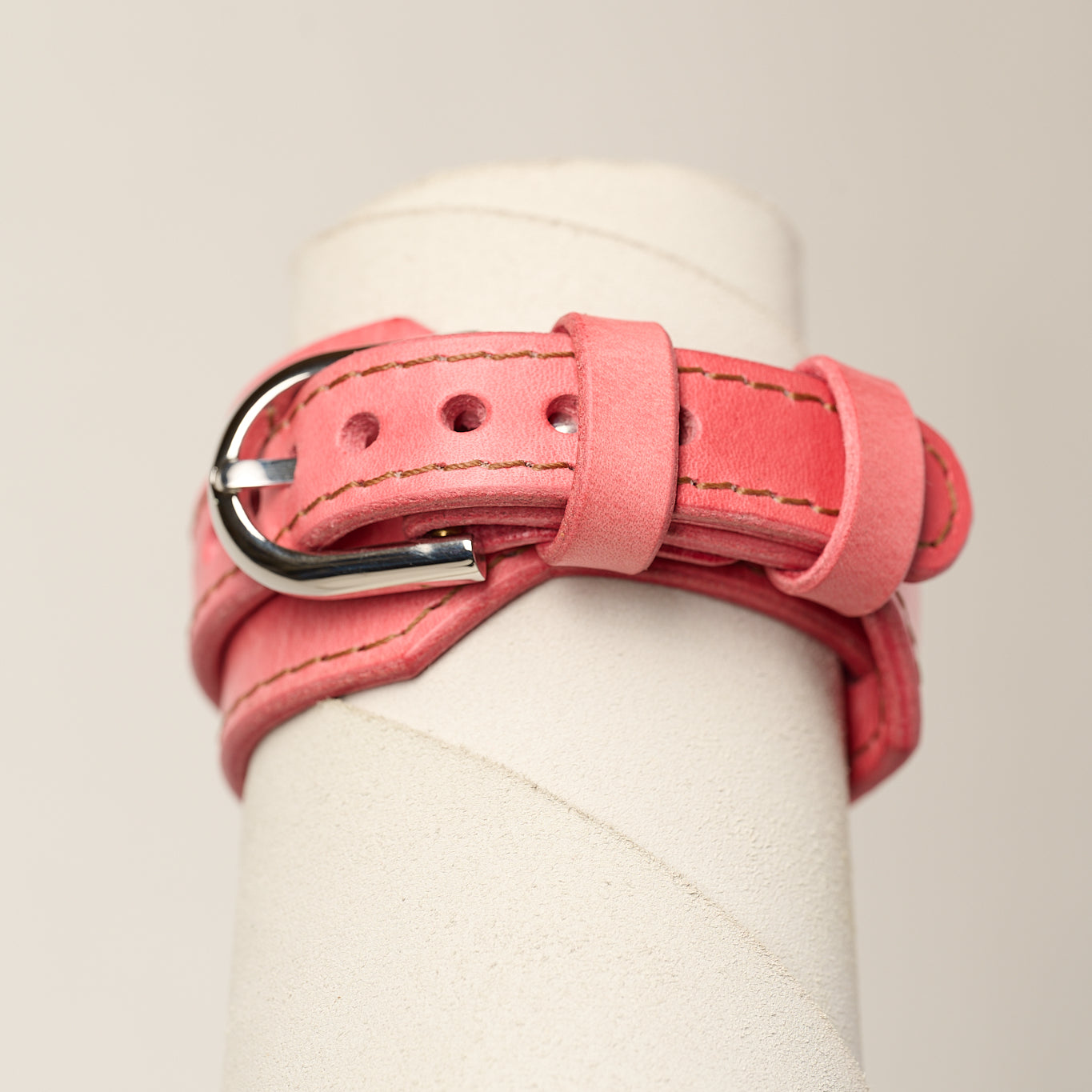 A pink leather wrap bracelet with a silver metal buckle, displayed on a white cylindrical mannequin stand against a light beige background.