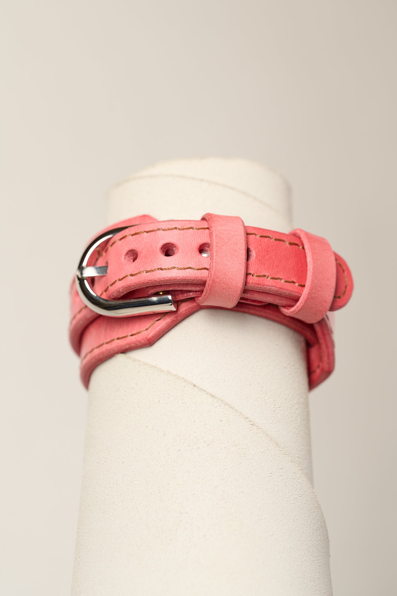 A pink leather wrap bracelet with a silver metal buckle, displayed on a white cylindrical mannequin stand against a light beige background.