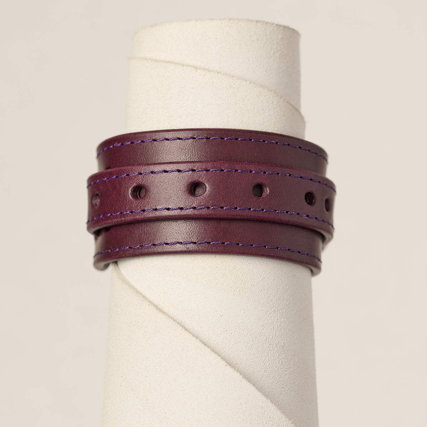 A dark purple leather wrap bracelet with purple stitching, displayed on a white cylindrical jewelry stand against a light beige background.
