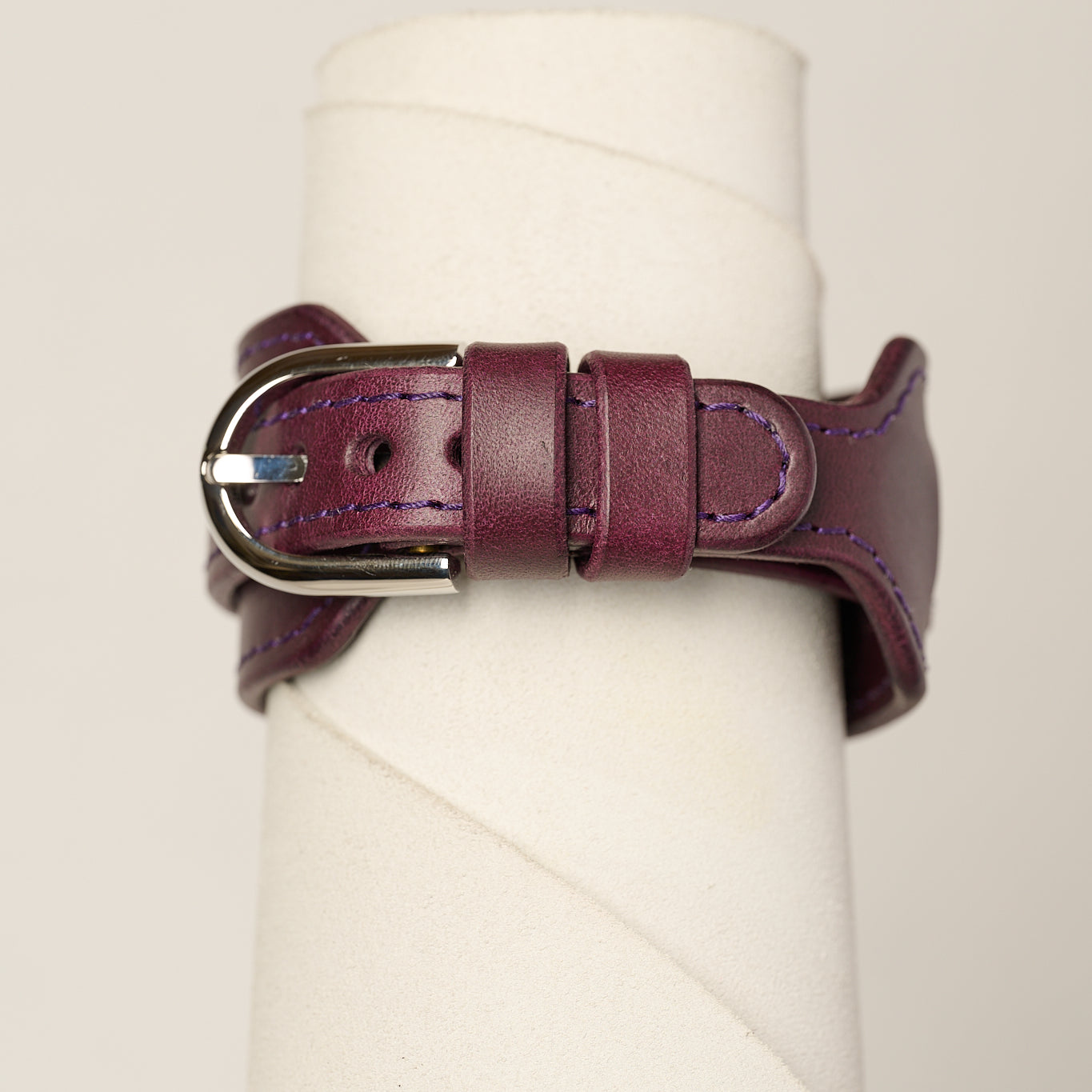 A dark purple leather watch strap with a silver-toned metal buckle, wrapped around a white cylindrical display stand.