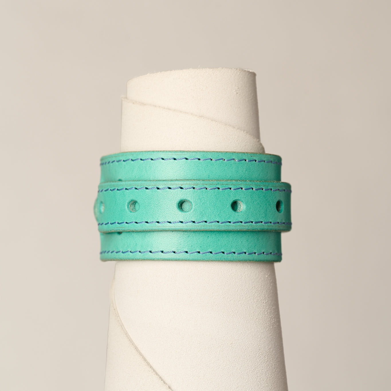 A turquoise leather wrap bracelet with matching stitching and metal eyelets, displayed on a white cone mannequin against a light gray background.