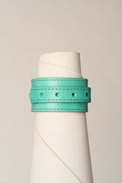 A turquoise leather wrap bracelet with matching stitching and metal eyelets, displayed on a white cone mannequin against a light gray background.