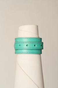 A turquoise leather wrap bracelet with matching stitching and metal eyelets, displayed on a white cone mannequin against a light gray background.