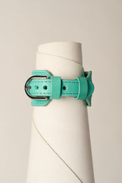 A mint green leather bracelet with a silver-toned metal buckle, displayed on a white cylindrical mannequin stand against a light beige background.