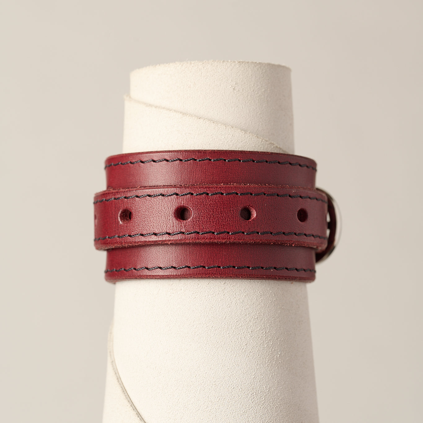 A burgundy leather wrap bracelet with dark contrast stitching and a silver buckle, displayed on a white textured cone stand against a light beige background.