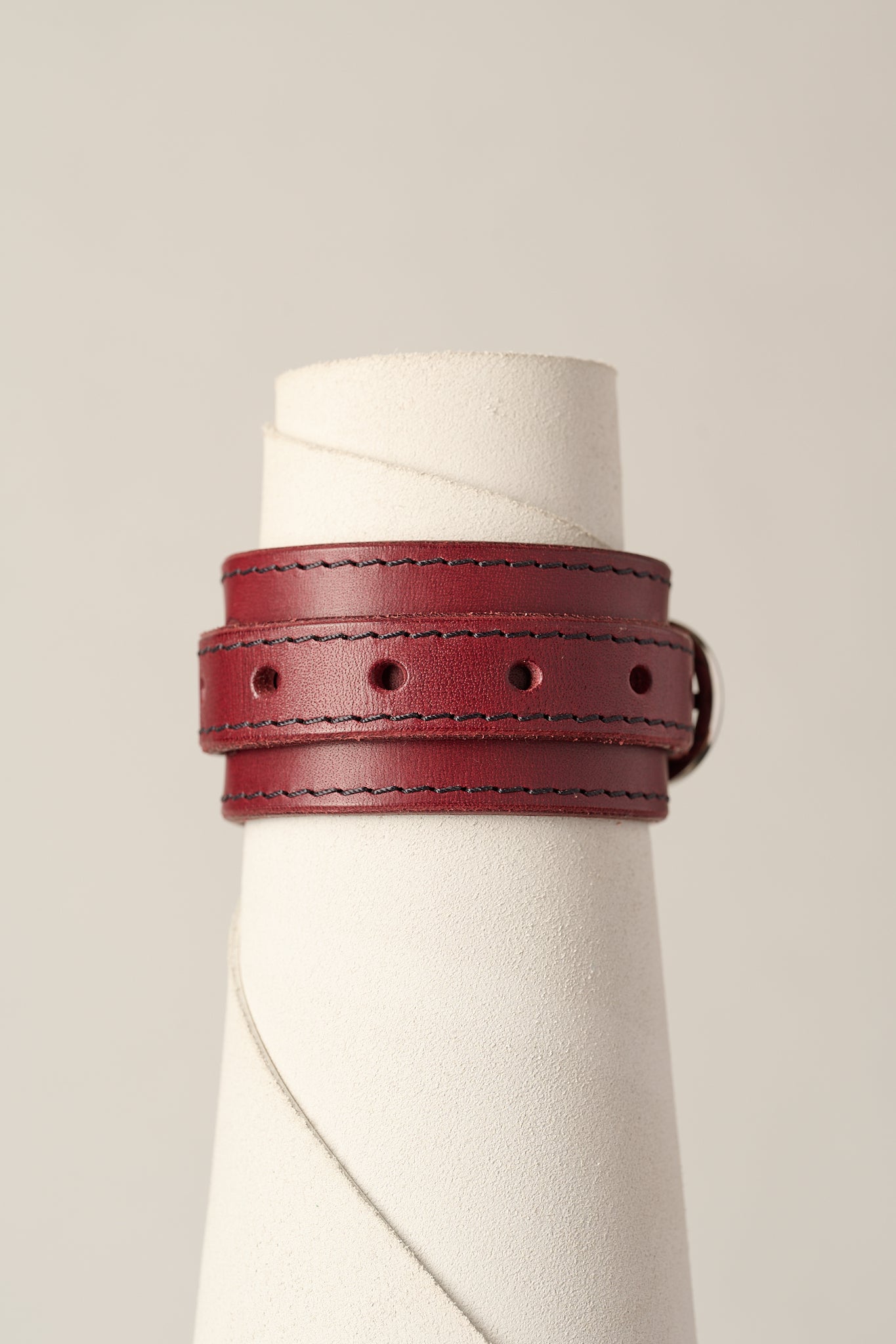 A burgundy leather wrap bracelet with dark contrast stitching and a silver buckle, displayed on a white textured cone stand against a light beige background.