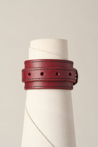 A burgundy leather wrap bracelet with dark contrast stitching and a silver buckle, displayed on a white textured cone stand against a light beige background.