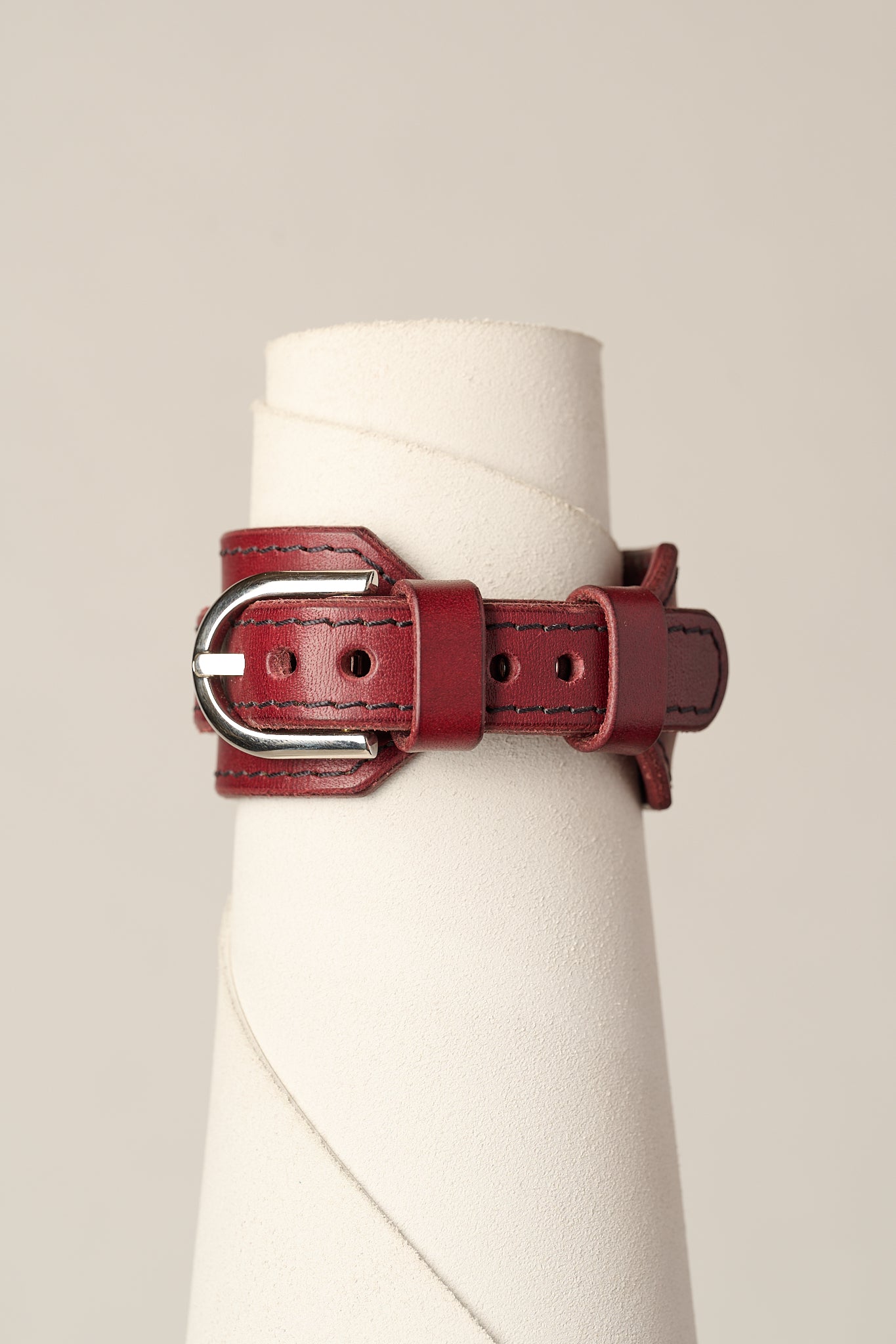 A dark red leather belt with a silver-tone metal buckle, displayed on a white fabric cone stand against a light beige background.