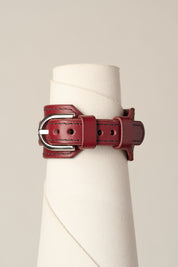 A dark red leather belt with a silver-tone metal buckle, displayed on a white fabric cone stand against a light beige background.