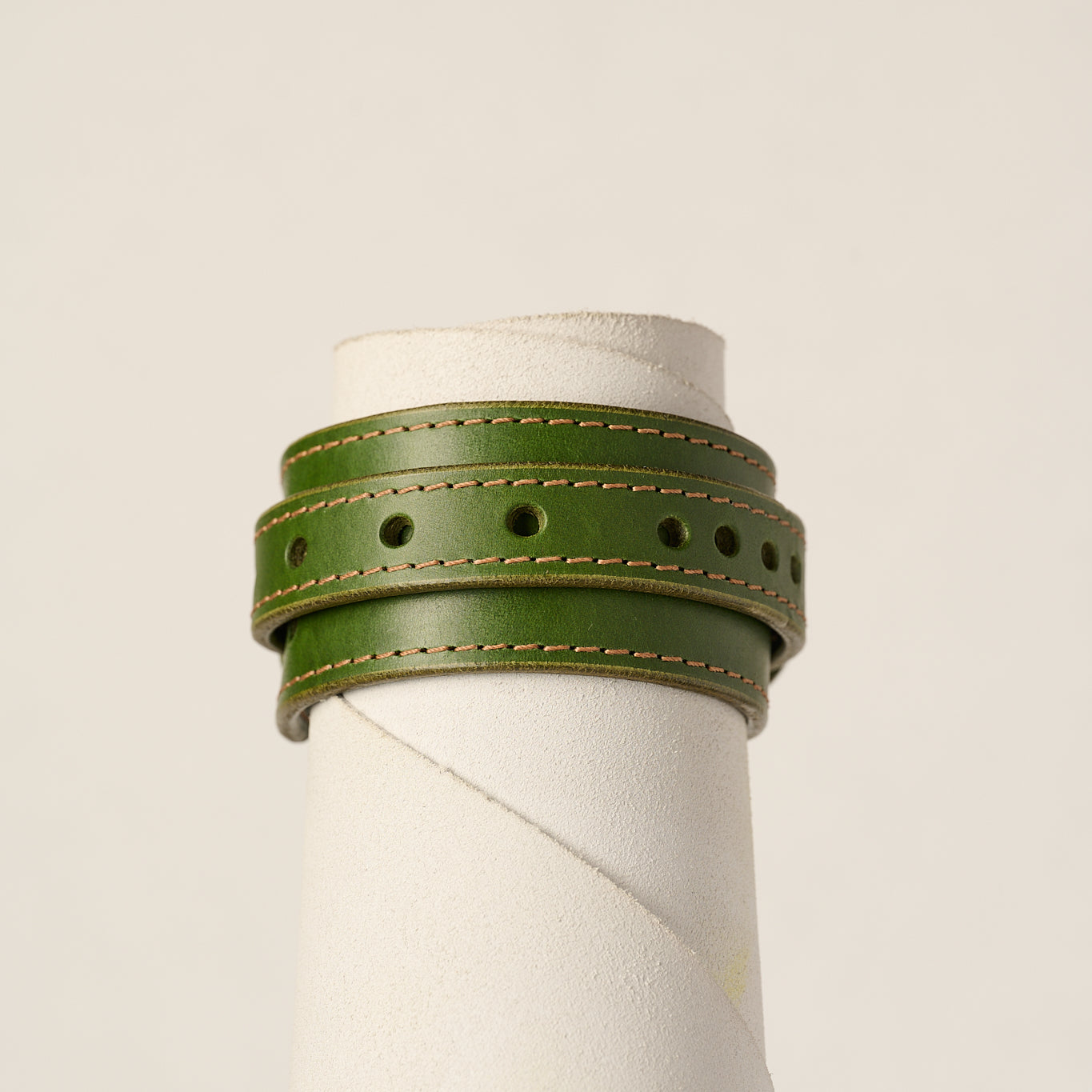 An olive green leather wrap bracelet with tan stitching, displayed on a white cylindrical mannequin stand against a light beige background.