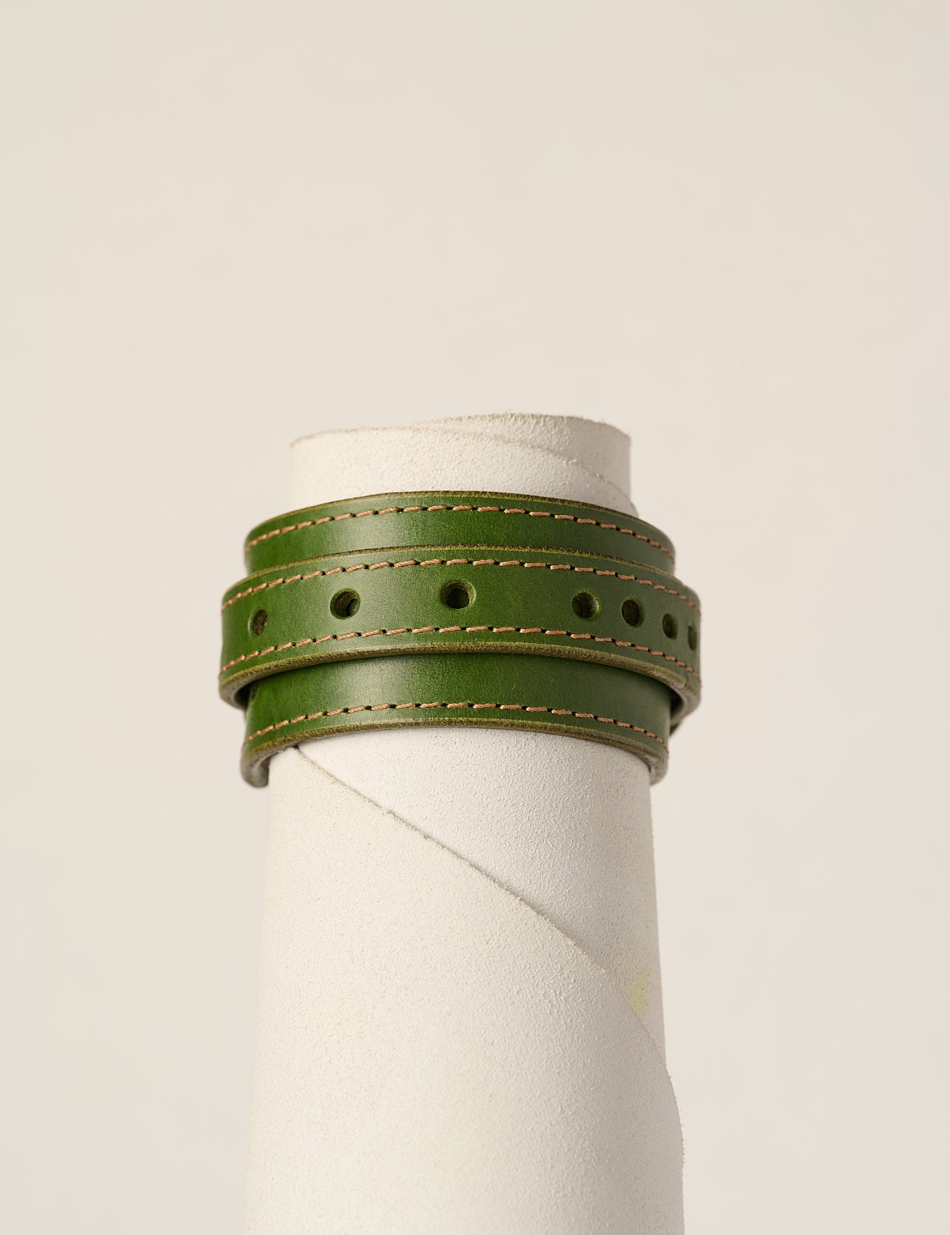 An olive green leather wrap bracelet with tan stitching, displayed on a white cylindrical mannequin stand against a light beige background.