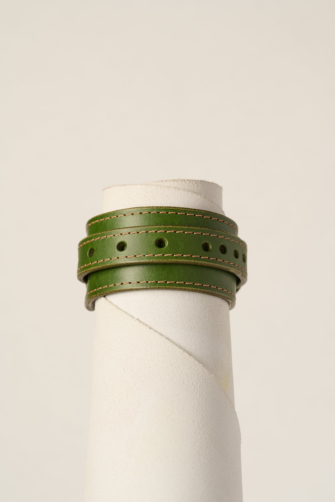 An olive green leather wrap bracelet with tan stitching, displayed on a white cylindrical mannequin stand against a light beige background.