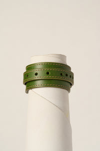 An olive green leather wrap bracelet with tan stitching, displayed on a white cylindrical mannequin stand against a light beige background.