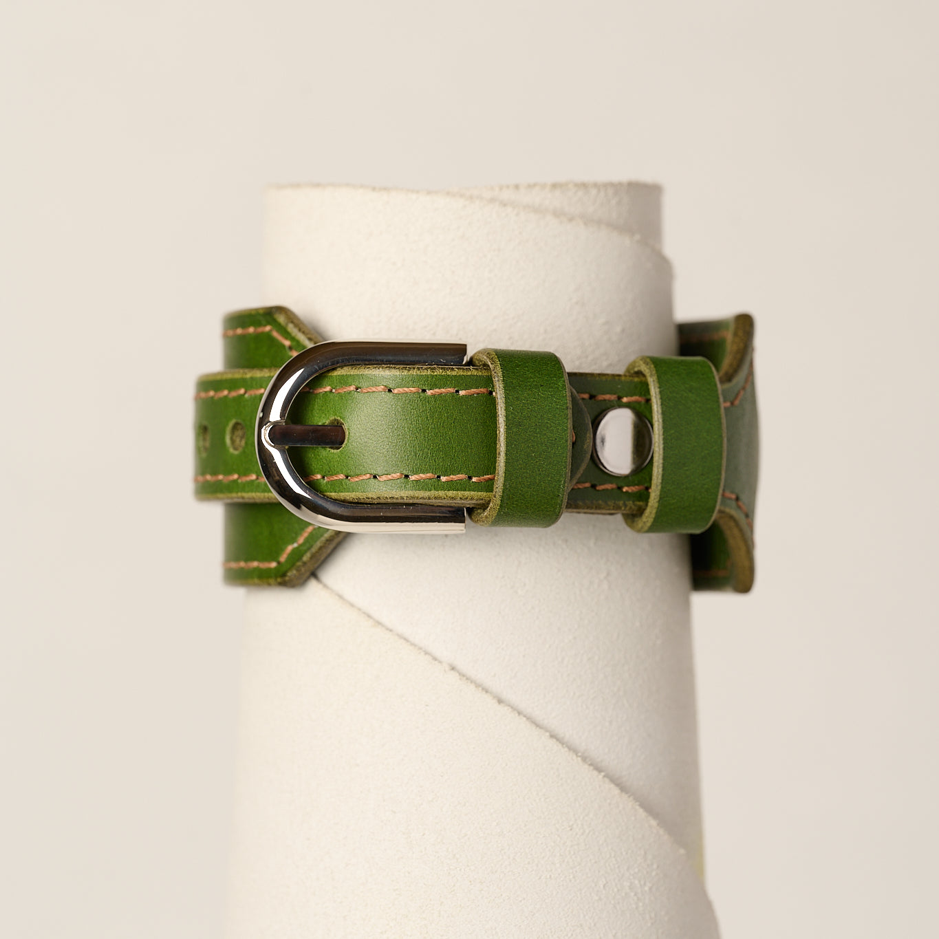 An olive green leather bracelet strap with a silver-toned metal buckle, displayed on a white cylindrical stand against a light beige background.
