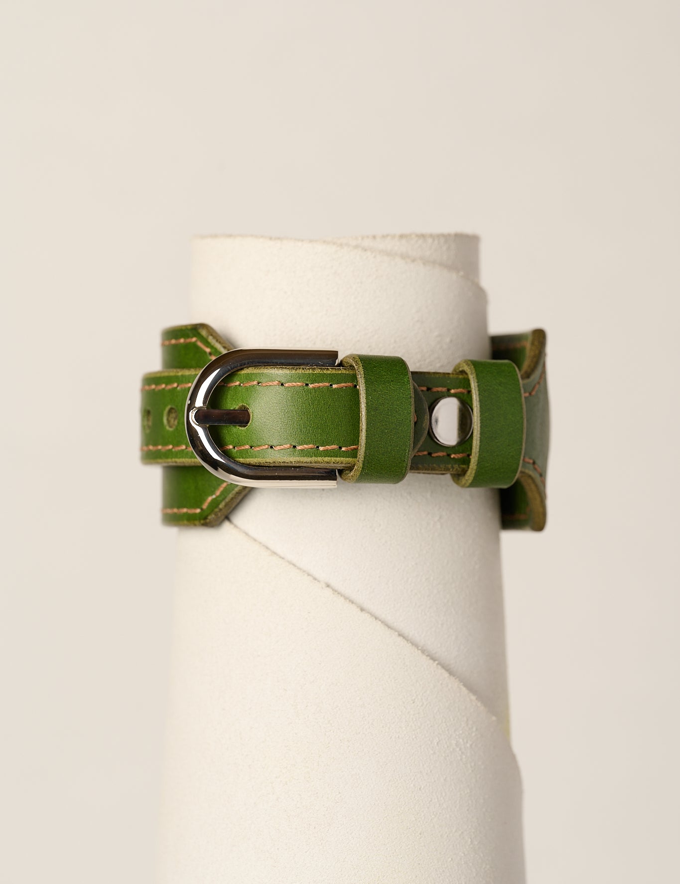 An olive green leather bracelet strap with a silver-toned metal buckle, displayed on a white cylindrical stand against a light beige background.