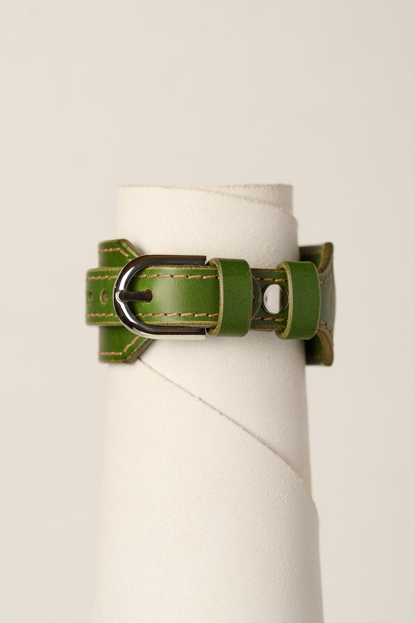 An olive green leather bracelet strap with a silver-toned metal buckle, displayed on a white cylindrical stand against a light beige background.