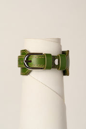 An olive green leather bracelet strap with a silver-toned metal buckle, displayed on a white cylindrical stand against a light beige background.