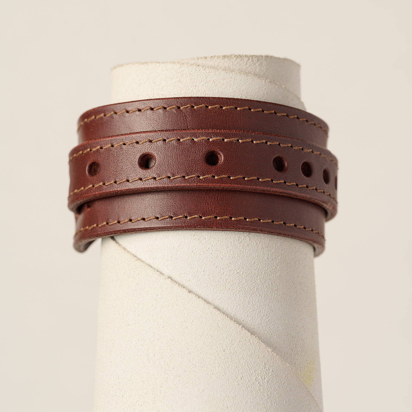 A brown leather wrap bracelet with double rows of stitching and circular cutouts, displayed on a textured white mannequin stand against a light beige background.