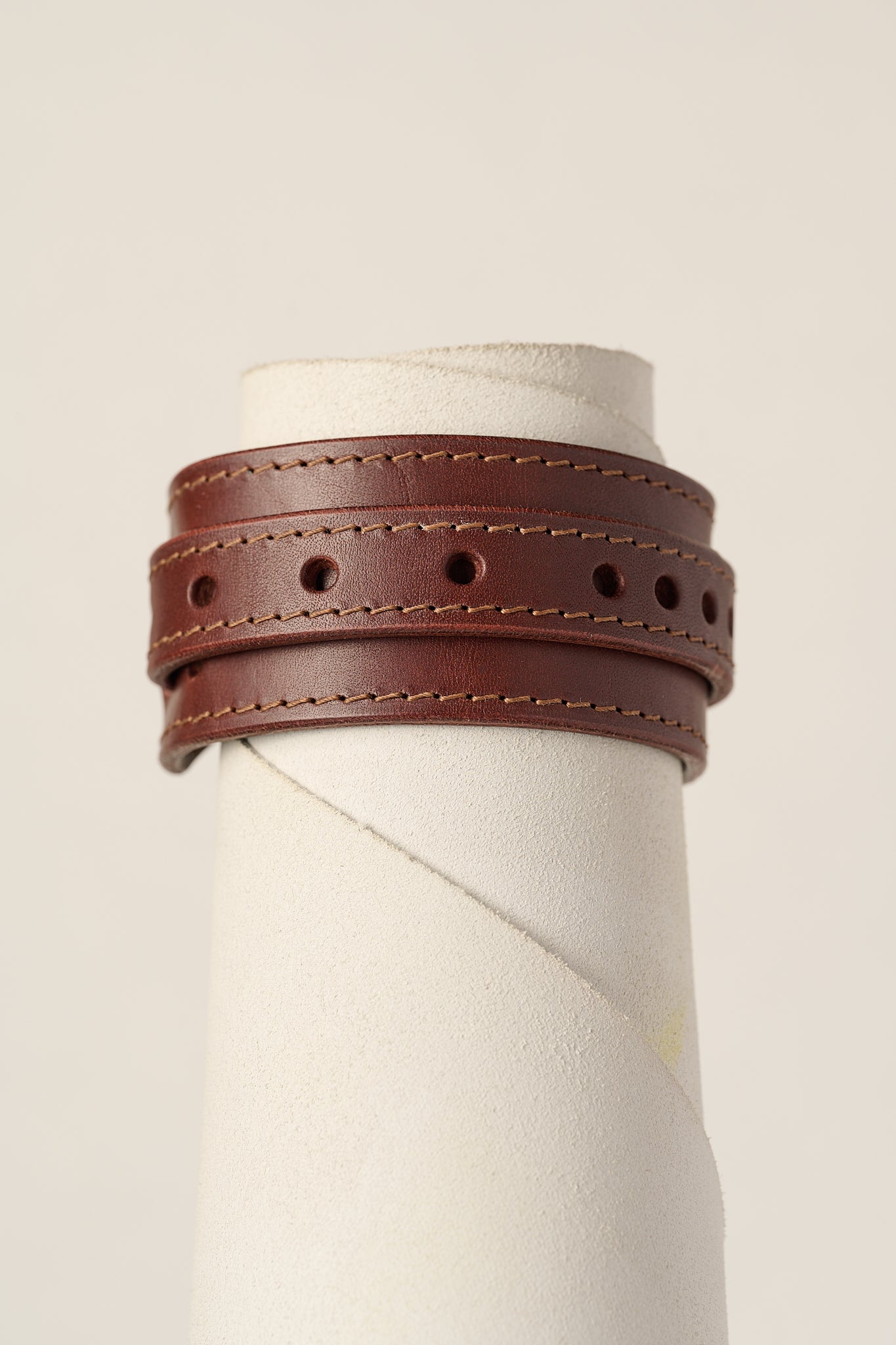 A brown leather wrap bracelet with double rows of stitching and circular cutouts, displayed on a textured white mannequin stand against a light beige background.