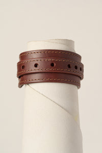 A brown leather wrap bracelet with double rows of stitching and circular cutouts, displayed on a textured white mannequin stand against a light beige background.