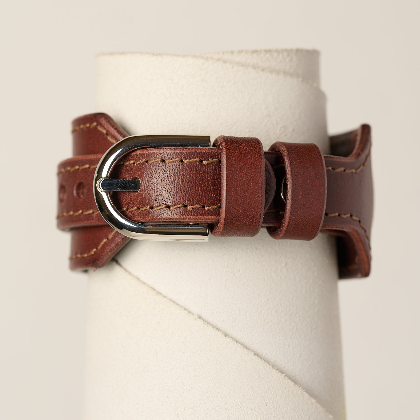 A brown leather bracelet strap with a silver-toned metal buckle, displayed on a white cylindrical stand against a light beige background.