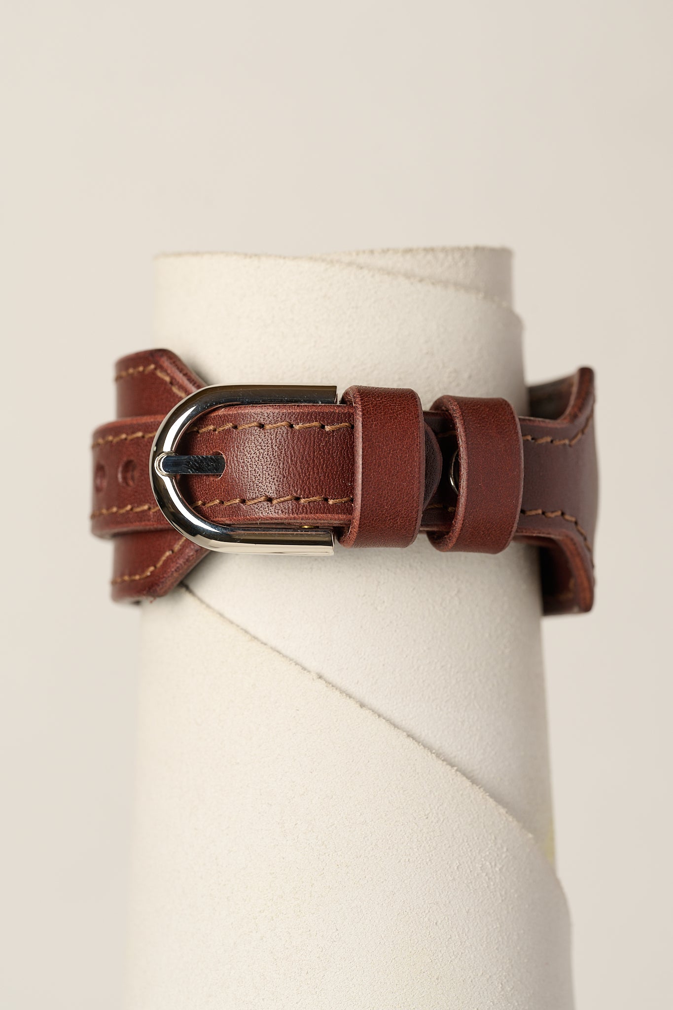 A brown leather bracelet strap with a silver-toned metal buckle, displayed on a white cylindrical stand against a light beige background.