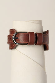 A brown leather bracelet strap with a silver-toned metal buckle, displayed on a white cylindrical stand against a light beige background.