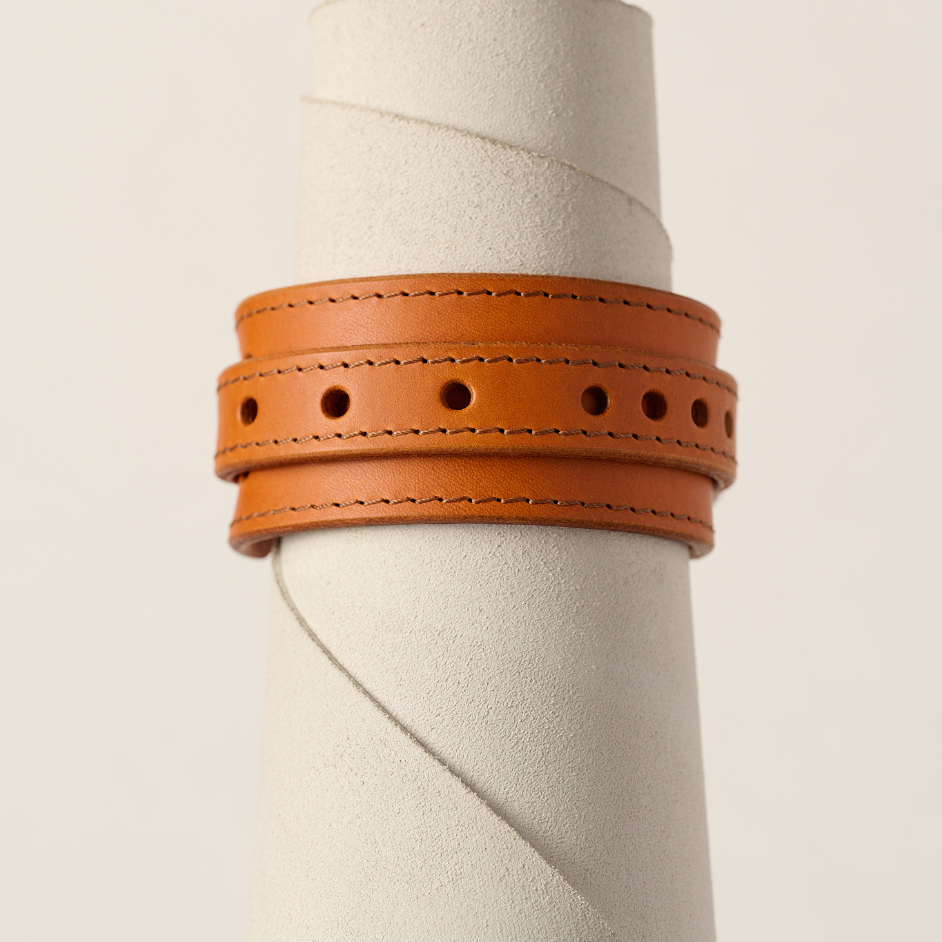 A orange leather wrap bracelet with double rows of stitching and circular cutouts, displayed on a textured white cylindrical stand against a light beige background.