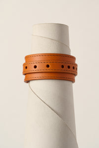 A orange leather wrap bracelet with double rows of stitching and circular cutouts, displayed on a textured white cylindrical stand against a light beige background.