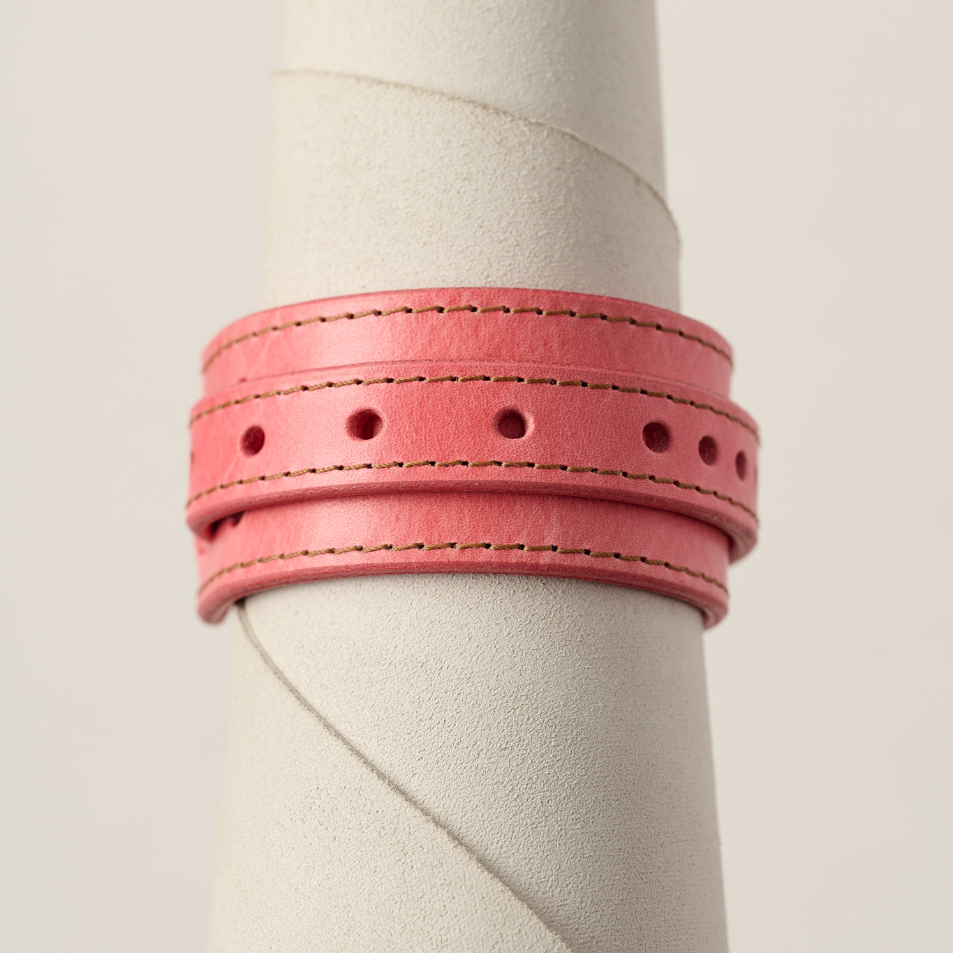 A pink leather bracelet with double stitched borders and circular cutouts, displayed on a textured off-white cylindrical stand against a light beige background.