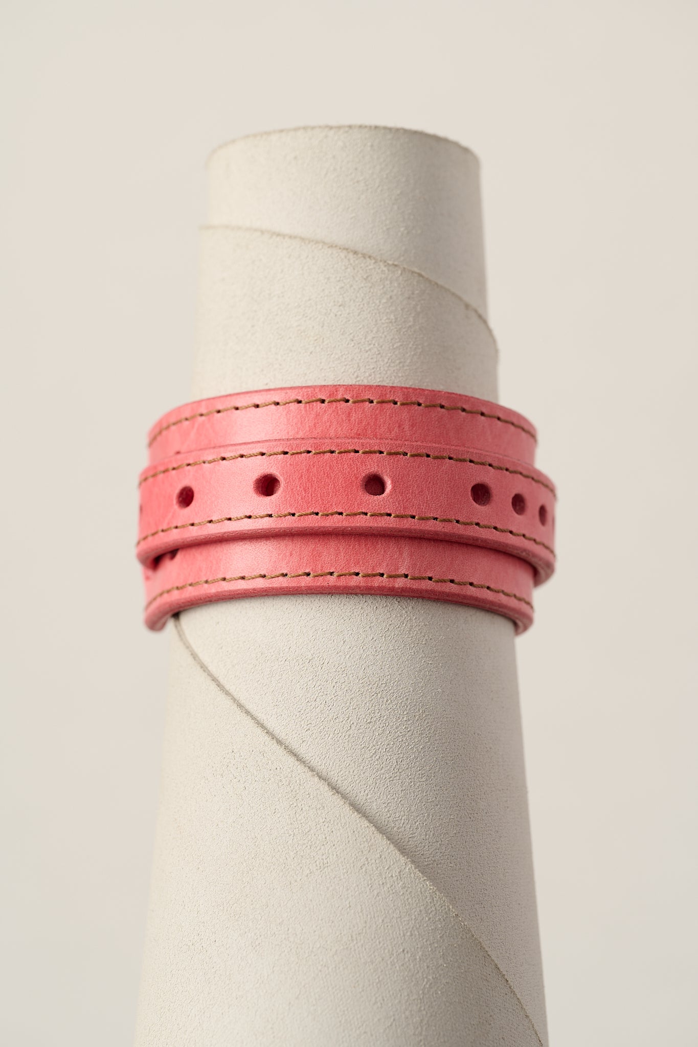 A pink leather bracelet with double stitched borders and circular cutouts, displayed on a textured off-white cylindrical stand against a light beige background.