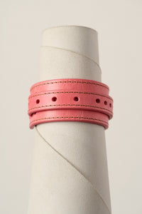 A pink leather bracelet with double stitched borders and circular cutouts, displayed on a textured off-white cylindrical stand against a light beige background.
