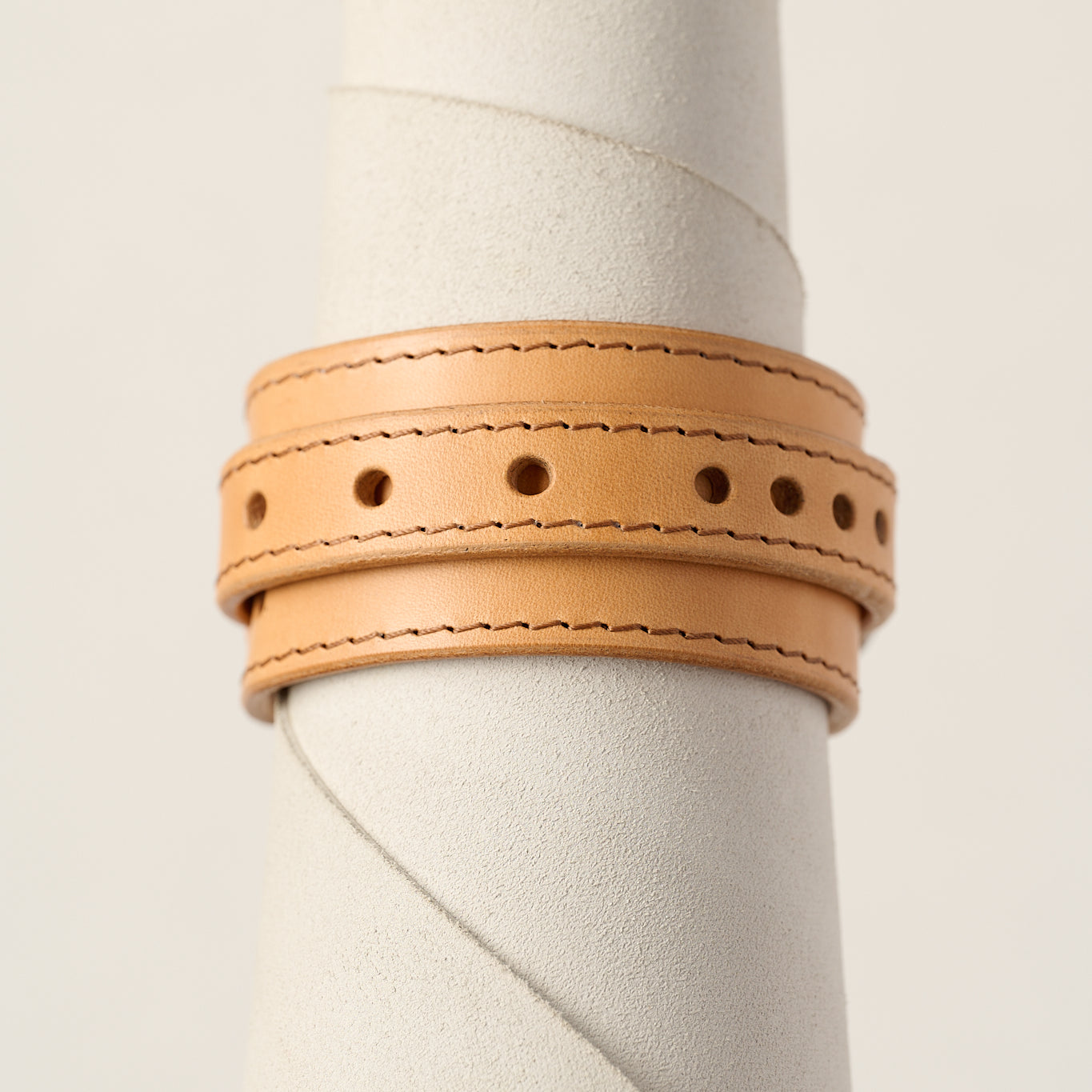 A light natural tan leather wrap bracelet with double rows of stitching and decorative punched holes, displayed on a textured white cylindrical stand.