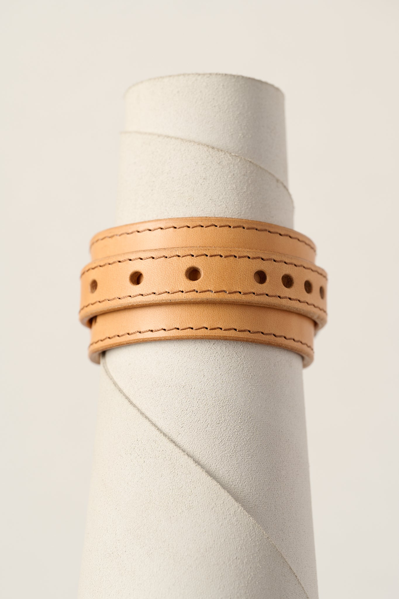 A light natural tan leather wrap bracelet with double rows of stitching and decorative punched holes, displayed on a textured white cylindrical stand.
