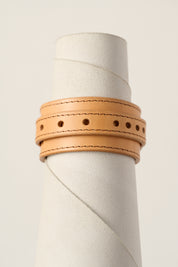 A light natural tan leather wrap bracelet with double rows of stitching and decorative punched holes, displayed on a textured white cylindrical stand.