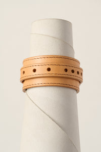 A light natural tan leather wrap bracelet with double rows of stitching and decorative punched holes, displayed on a textured white cylindrical stand.
