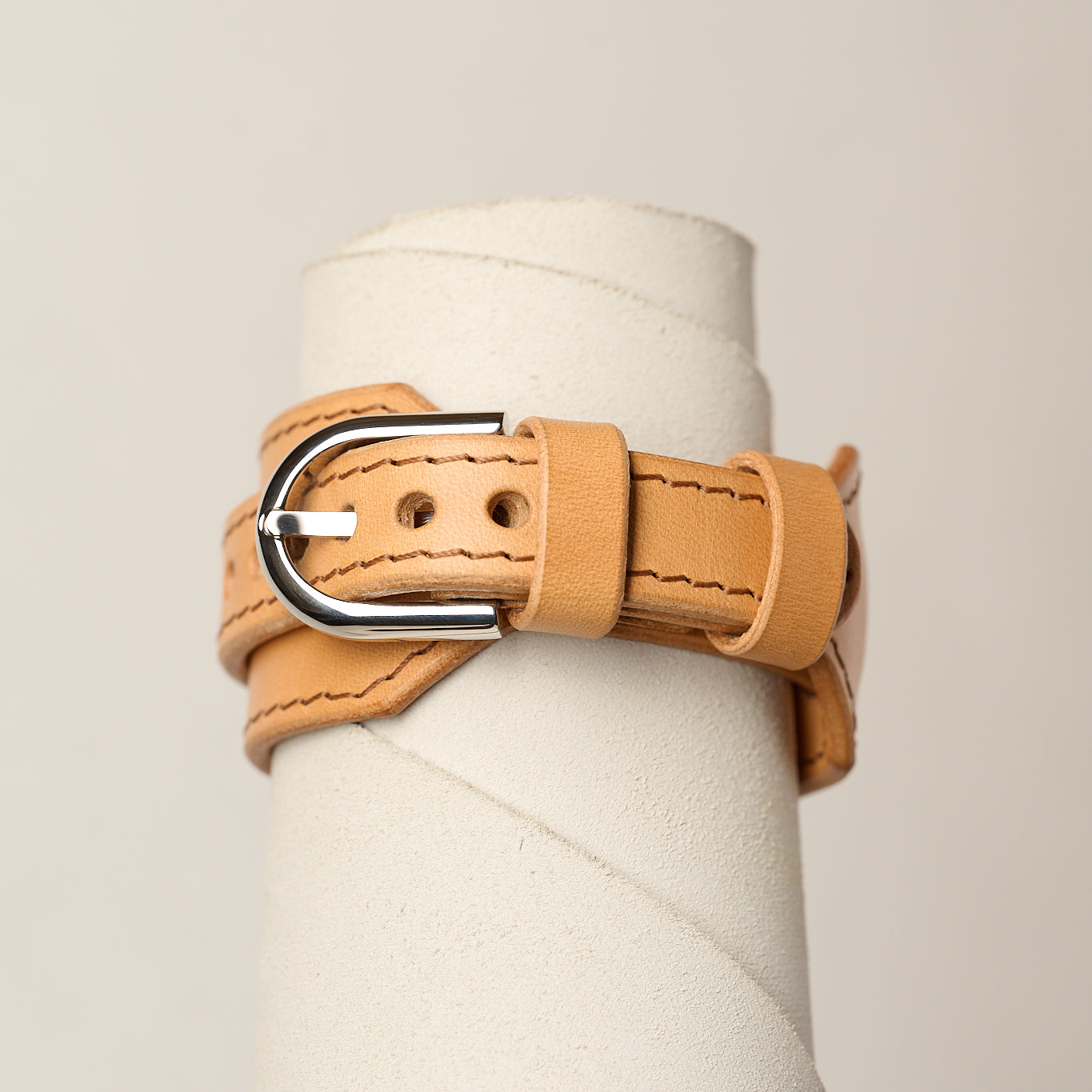 A light tan leather wrap bracelet with a silver-tone metal buckle, displayed on a white cylindrical mannequin stand against a light beige background.