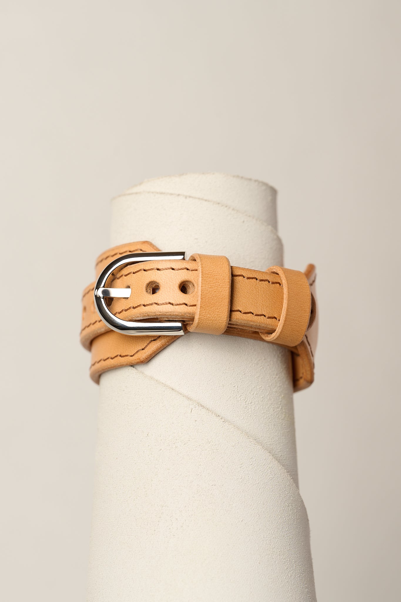 A light tan leather wrap bracelet with a silver-tone metal buckle, displayed on a white cylindrical mannequin stand against a light beige background.