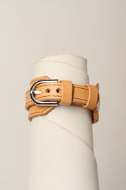 A light tan leather wrap bracelet with a silver-tone metal buckle, displayed on a white cylindrical mannequin stand against a light beige background.