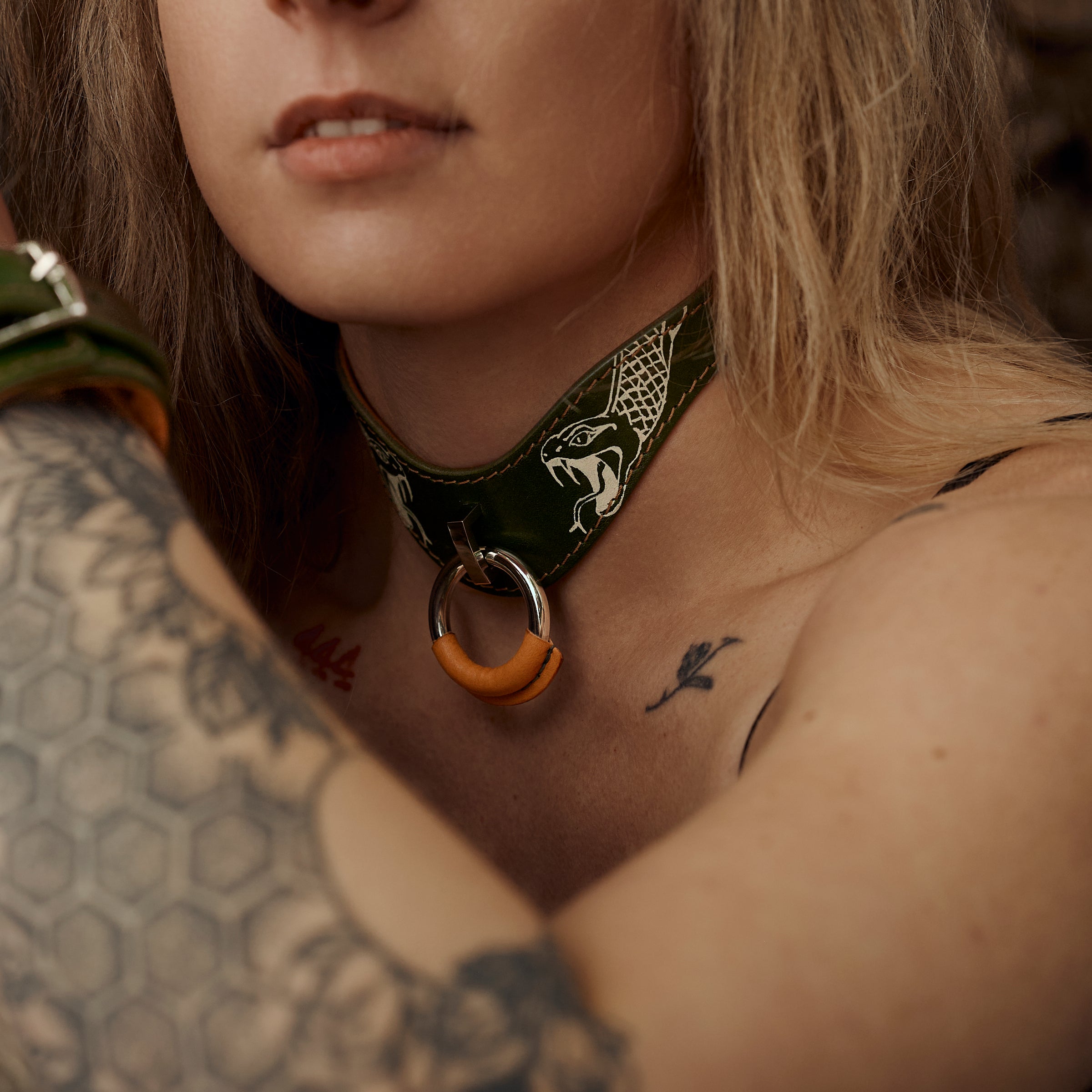 A close-up portrait of a woman with blonde hair and visible tattoos wearing a dark green tattooed leather choker necklace with a circular orange and silver pendant.