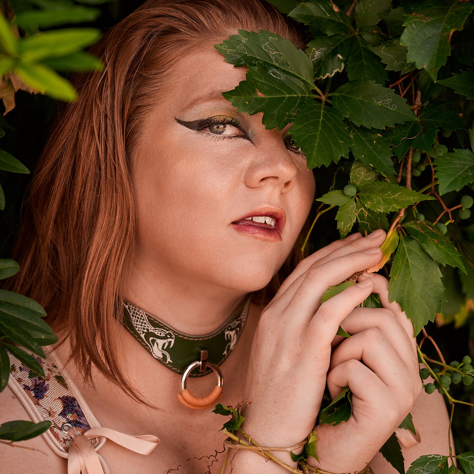 Woman with red hair and dark makeup holding green leaves, surrounded by foliage wearing a leather necklace tattooed with a snake 